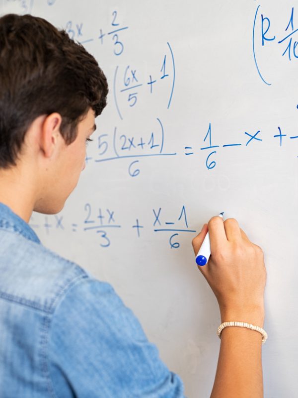 Back view of high school student solving math problem on whiteboard in classroom. Young man writing math solution on white board using marker. College guy solving math expression during lesson.